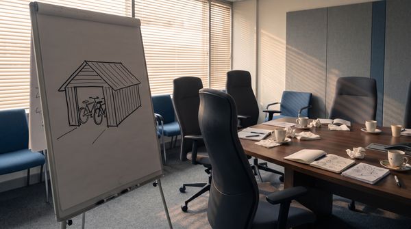 Empty corporate meeting room after a long meeting, with a flip chart showing a simple marker sketch of a bicycle shed beside a conference table strewn with coffee cups and notepads