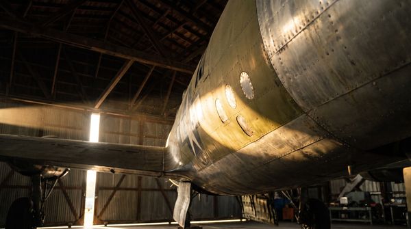 Underside of a vintage WWII bomber fuselage in a dim hangar, riveted aluminium skin with patched panels catching a shaft of warm light
